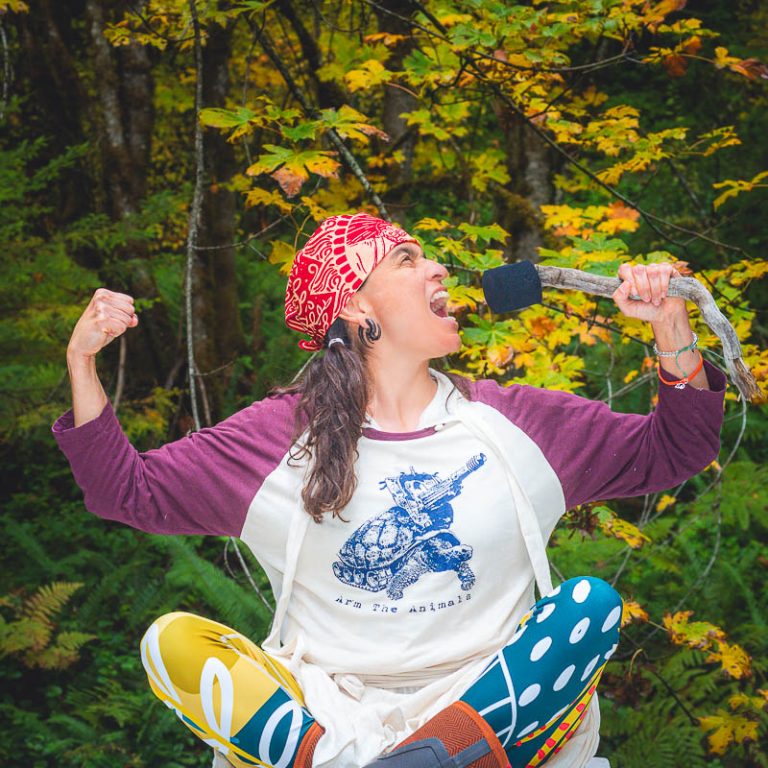 A woman in the forest shouting into a microphone with her arms raised and fist clenched